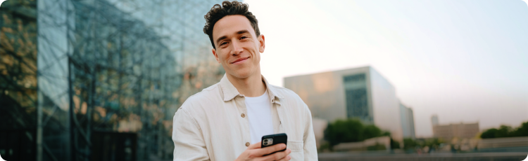 MÅody mÄÅ¼czyzna z uÅmiechem trzyma smartfon na tle nowoczesnych budynkÃ³w â symbol Åwiadomego podejÅcia do finansÃ³w i rozpoczÄcia oszczÄdzania na emeryturÄ z IKZE