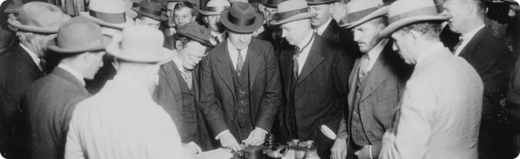 Photo historique en noir et blanc de nÃ©gociants en costume et chapeau rÃ©unis autour dâune table de nÃ©gociation au Chicago Board of Trade, illustrant les dÃ©buts de la spÃ©culation sur les matiÃ¨res premiÃ¨res et de la nÃ©gociation sur le marchÃ©
