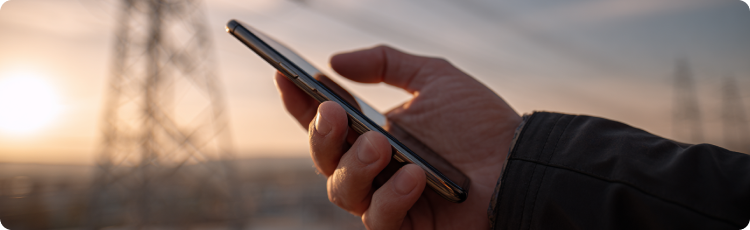 Tampak dekat tangan memegang smartphone dengan latar belakang jaringan listrik di waktu senja â melambangkan manajemen energi digital, teknologi smart grid, dan investasi di infrastruktur utilitas.
