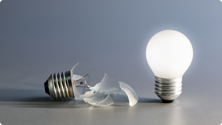 Gambar dua bohlam rusak dengan kaca pecah dan filamen yang putus pada latar netral