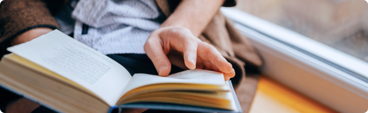 Close-up seseorang memegang dan membaca buku di dekat jendela, melambangkan proses belajar finansial dan pengetahuan investasi