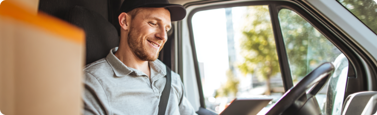 Pengemudi pengiriman tersenyum mengenakan kaus polo abu-abu dan topi, duduk di dalam van dengan kotak kardus terlihat di bagian depan, sambil memeriksa perangkat genggam.