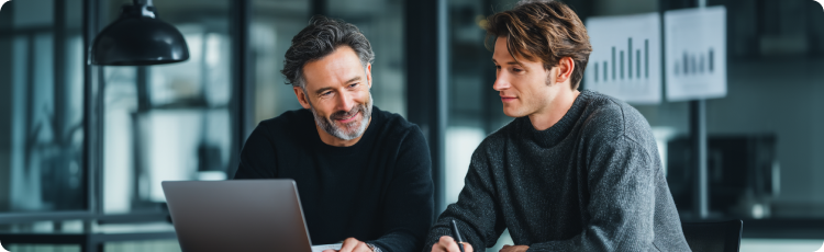 Dois homens sentados Ã  mesa, envolvidos numa discussÃ£o sobre investimentos enquanto utilizam um computador portÃ¡til