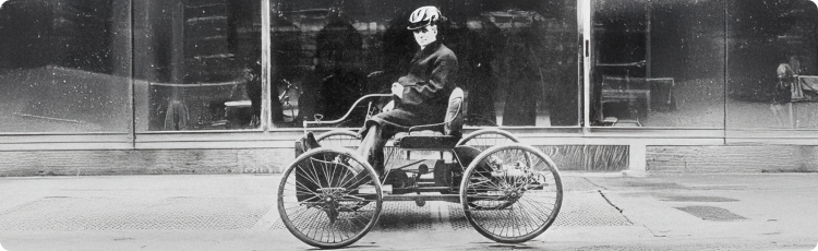 Histórico automóvil de tres ruedas conducido por una persona en una calle de la ciudad, que destaca las primeras innovaciones de ingenierÃa relevantes para el desarrollo industrial y económico a largo plazo