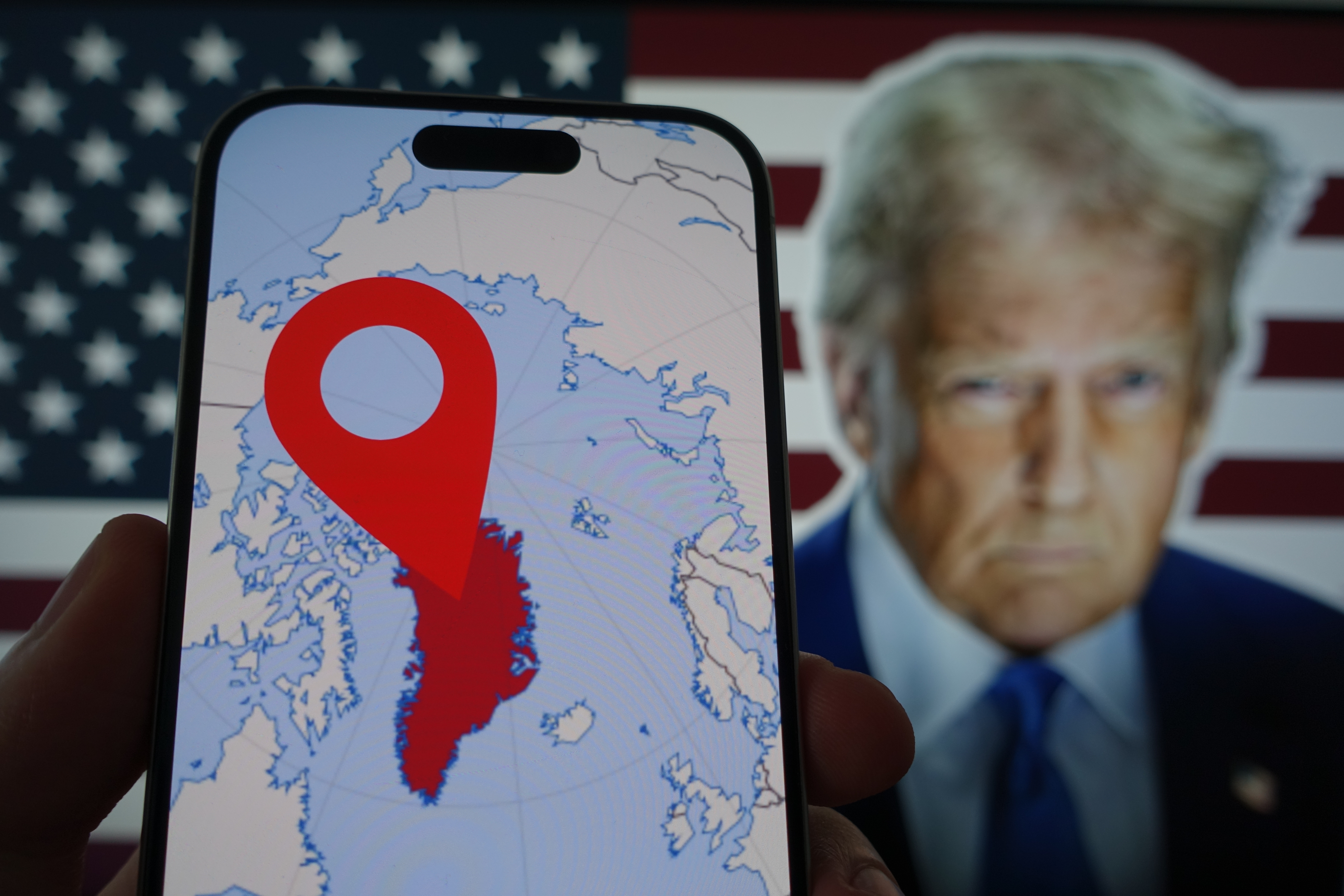 Kiel, SH, Germany March 29 2025: Smartphone Displaying a map of Geenland in Front of Blurred Image of Donald Trump and American Flag