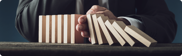 Seorang pria mengenakan setelan jas sedang menahan balok kayu seperti domino agar tidak jatuh, merepresentasikan profesionalisme dan manajemen risiko