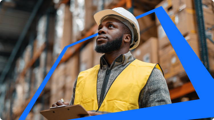 Raktári dolgozó fehér védÅsisakban és sárga láthatósági mellényben, kezében csiptetÅs mappával, felfelé néz a magas polcok között, amelyek dobozokkal vannak tele.