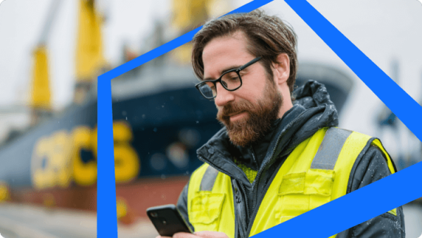 พนักงานท่าเรือสวมเซฟตี้ ใช้สมาร์ตโฟนหน้าเรือบรรทุกสินค้า แสดงการติดตามโลจิสติกส์และการลงทุนเรียลไทม์