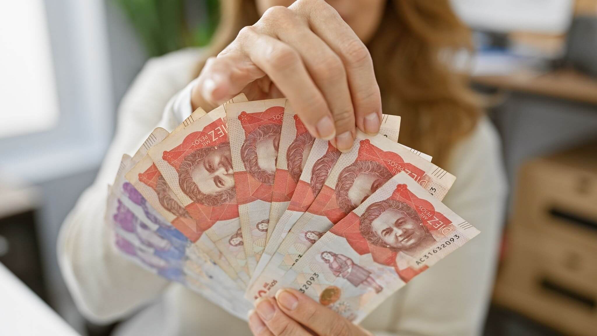Joven hispana sostiene pesos colombianos en un ambiente de oficina bien iluminado, sugiriendo negocios y finanzas.