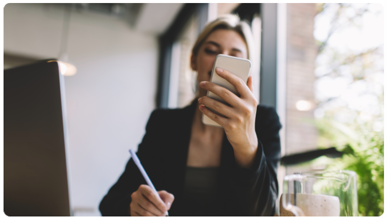 Une femme assise Ã  un bureau, utilisant son smartphone pour prendre des notes.